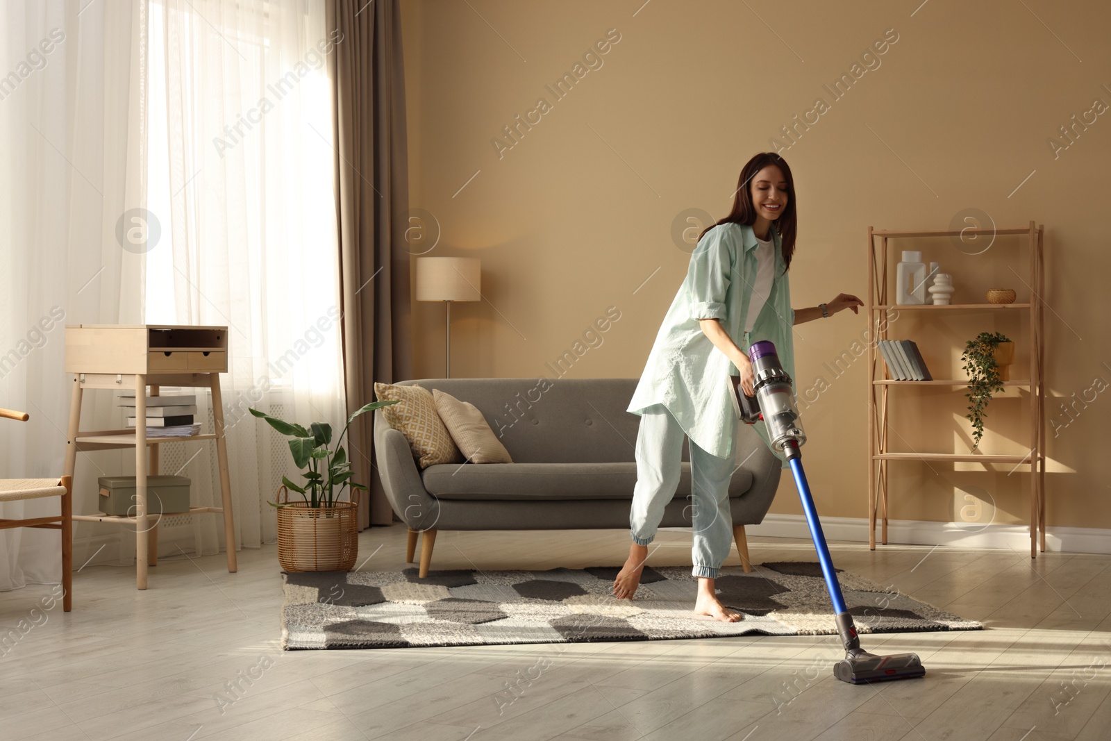 Photo of Smiling young woman cleaning rug with cordless vacuum cleaner in living room