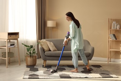 Photo of Smiling young woman cleaning rug with cordless vacuum cleaner in living room