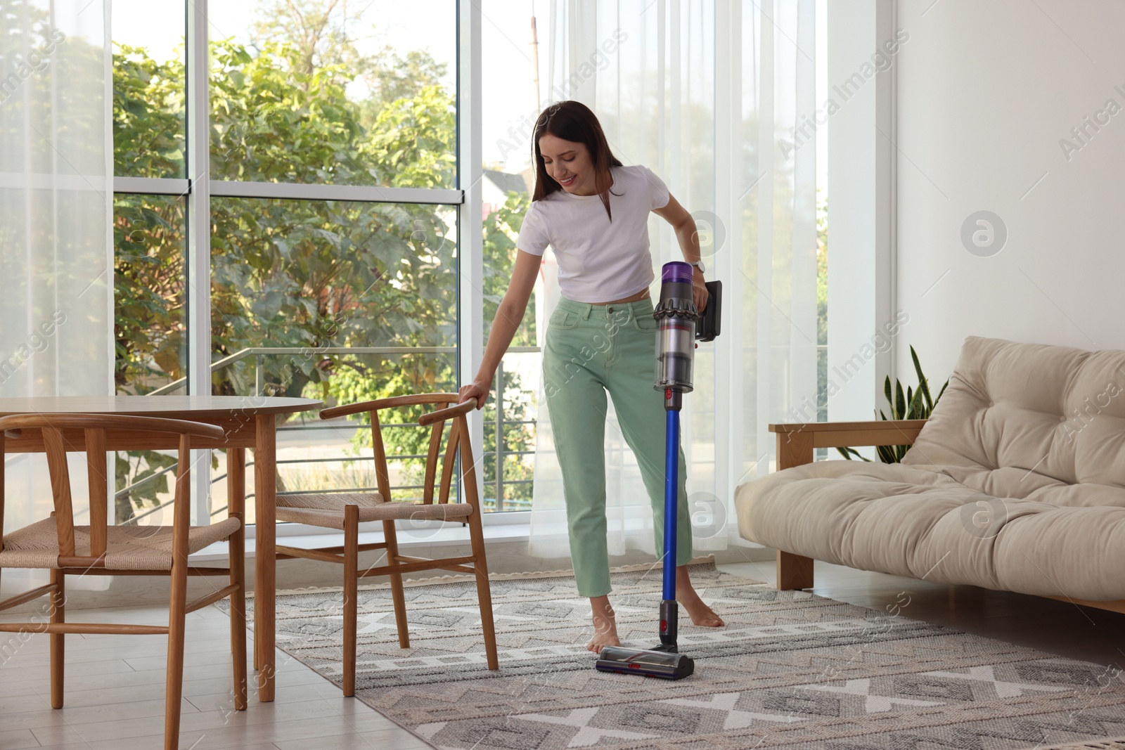 Photo of Smiling young woman cleaning rug with cordless vacuum cleaner at home