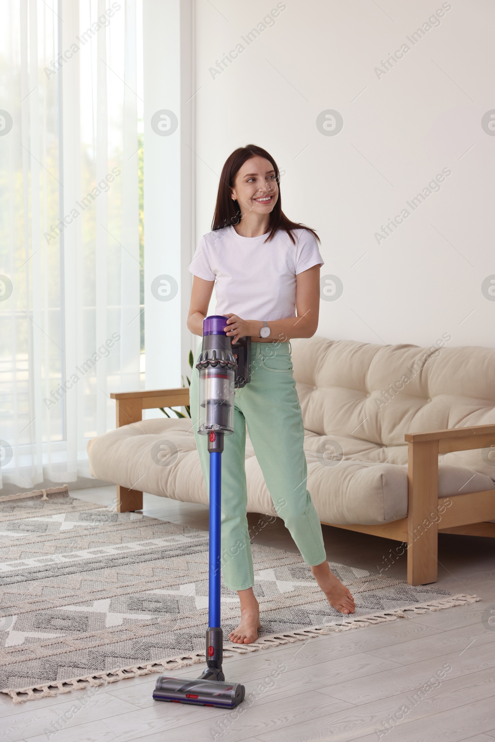 Photo of Smiling young woman cleaning floor with cordless vacuum cleaner at home