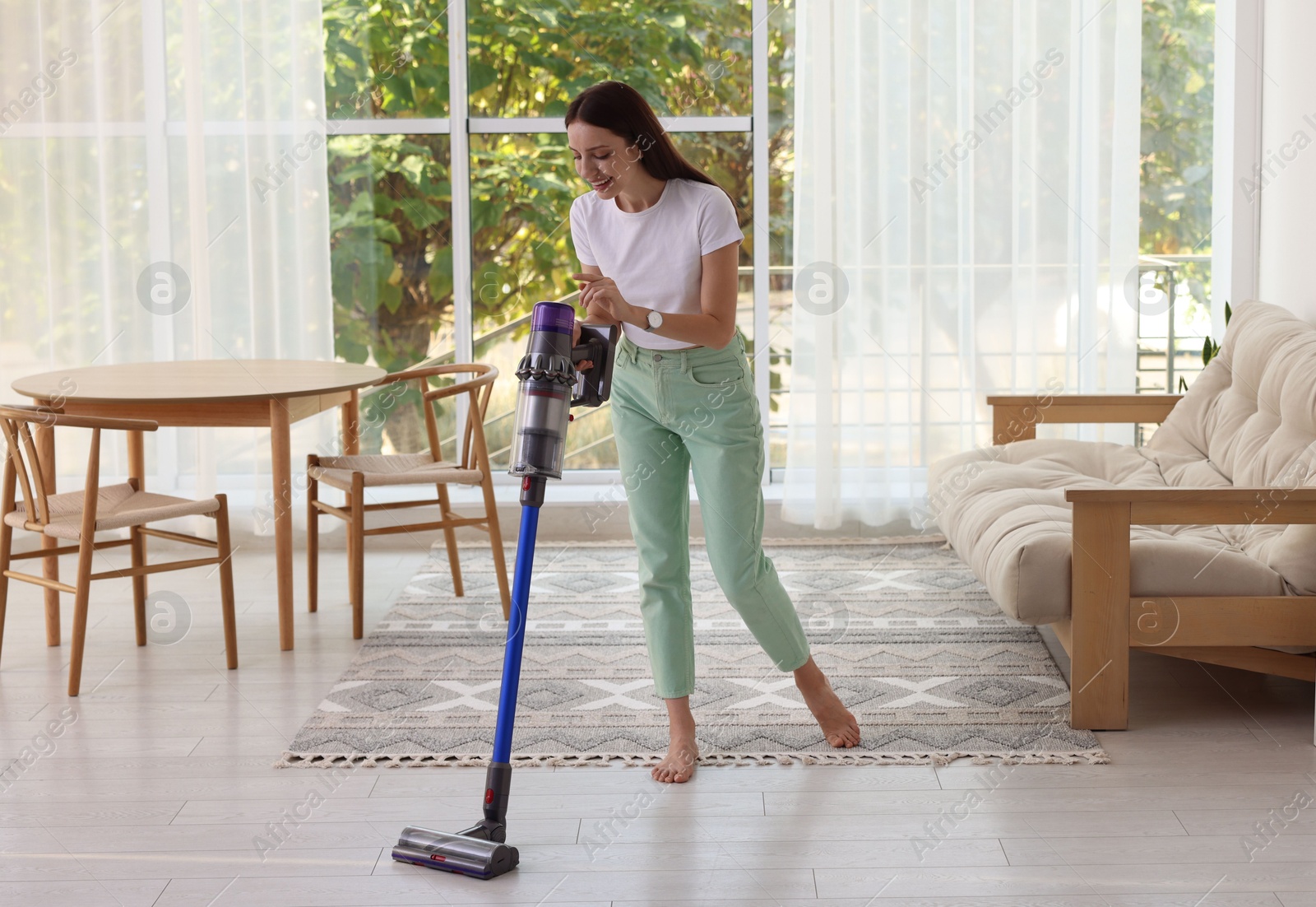 Photo of Smiling young woman cleaning floor with cordless vacuum cleaner at home