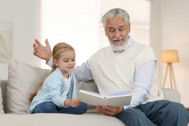 Photo of Grandpa and his granddaughter reading book together on sofa at home