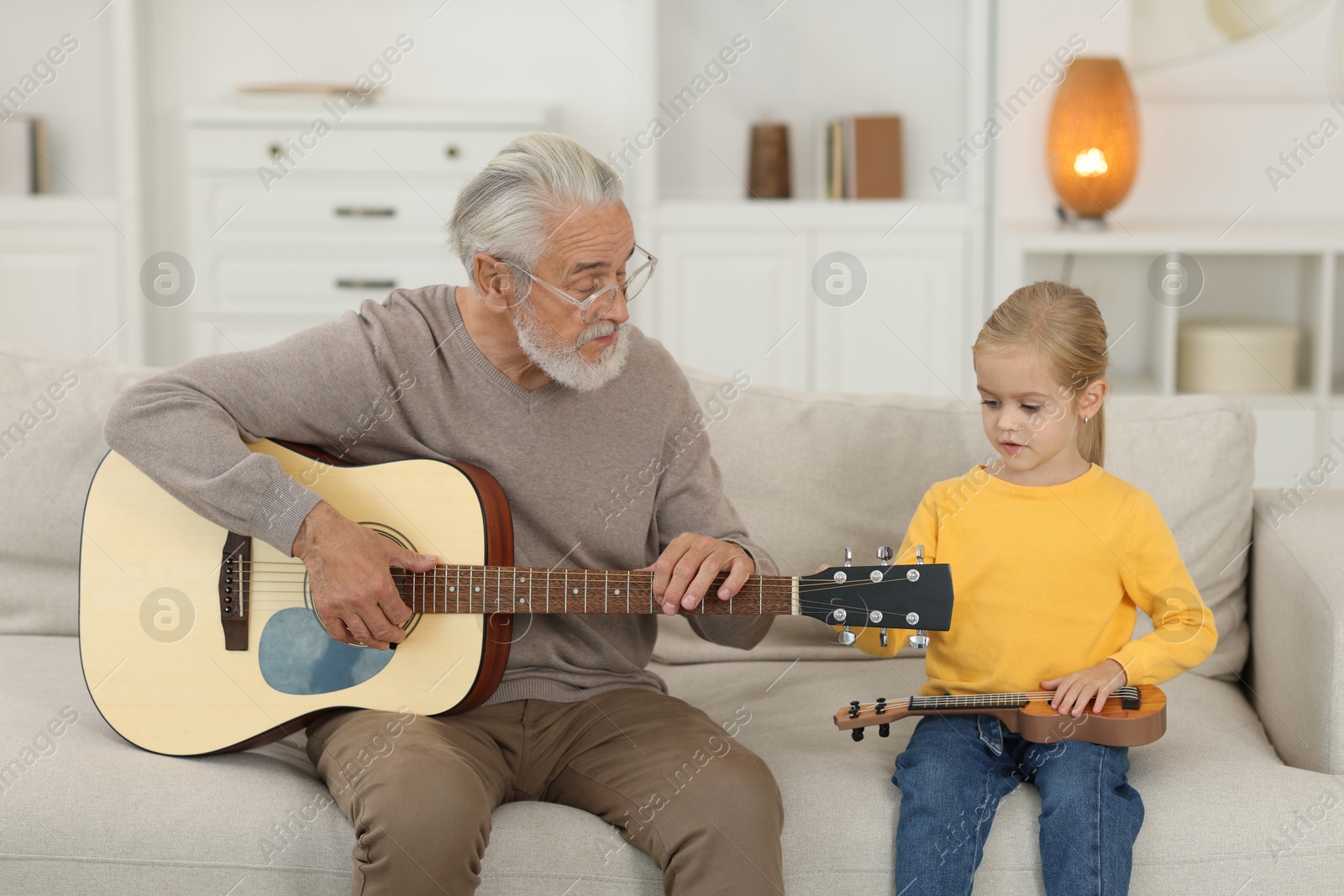 Photo of Grandpa teaching his granddaughter to play guitar on sofa at home