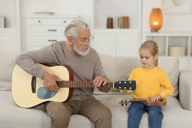 Photo of Grandpa teaching his granddaughter to play guitar on sofa at home