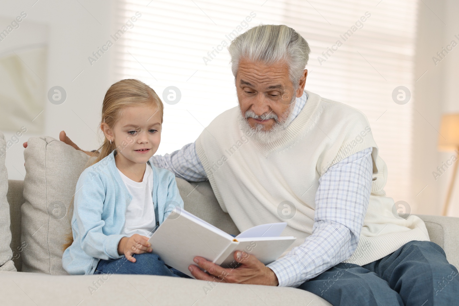 Photo of Grandpa and his granddaughter reading book together on sofa at home