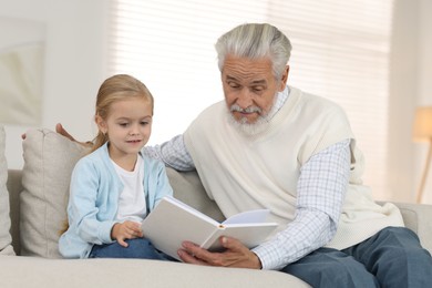 Photo of Grandpa and his granddaughter reading book together on sofa at home