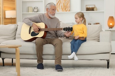 Photo of Grandpa teaching his granddaughter to play guitar on sofa at home