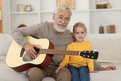 Photo of Grandpa teaching his granddaughter to play guitar on sofa at home