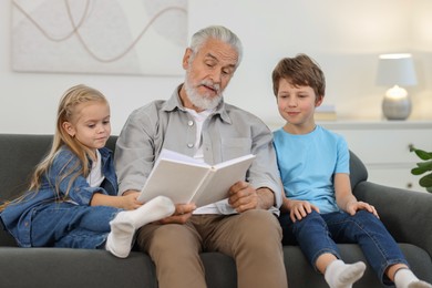 Photo of Grandpa and his grandkids reading book together on sofa at home