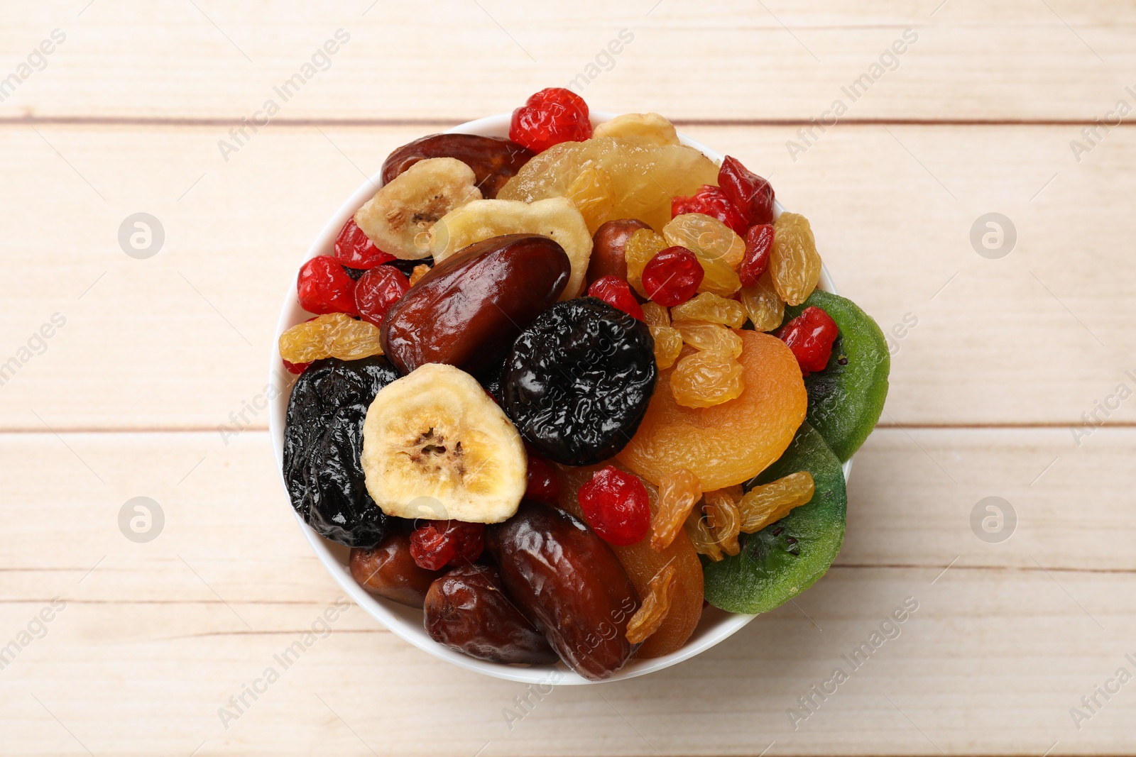 Photo of Mix of different dried fruits in bowl on white wooden table, top view