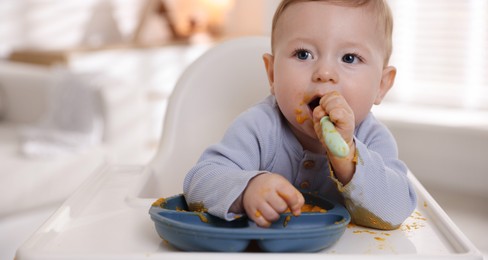 Photo of Cute little baby eating healthy food in high chair indoors