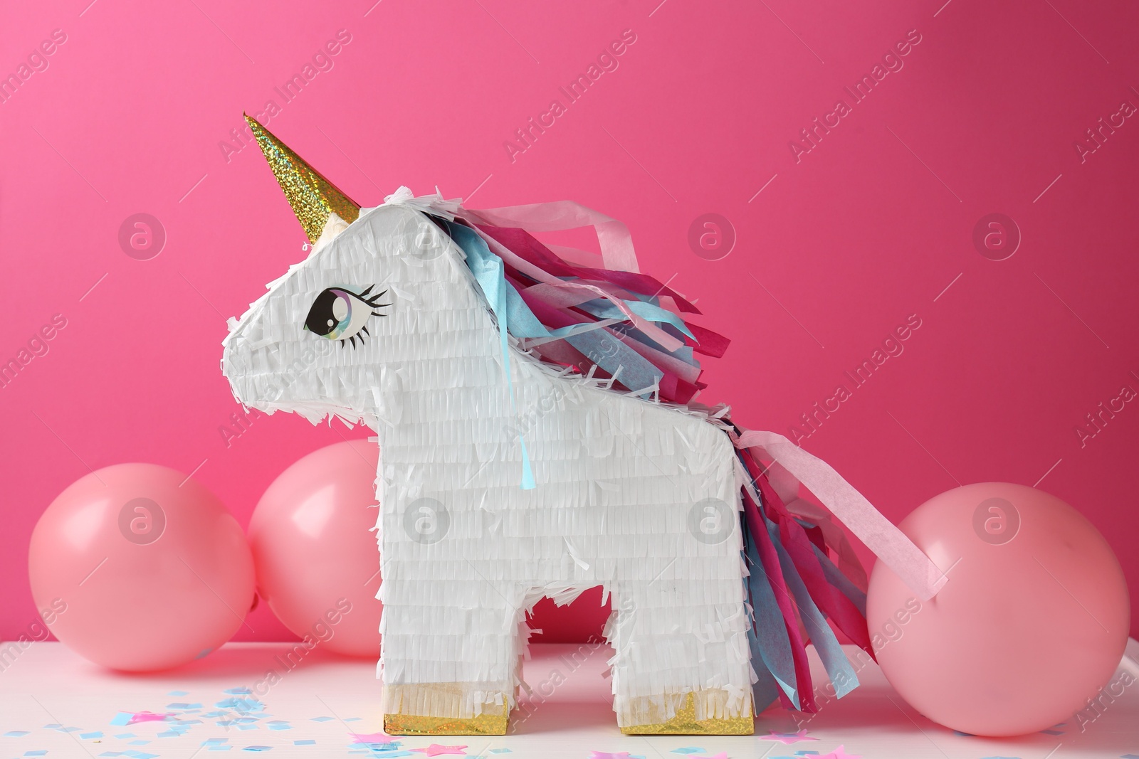 Photo of Bright pinata in shape of unicorn and balloons on white table against pink background