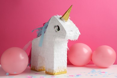 Photo of Bright pinata in shape of unicorn and balloons on white table against pink background