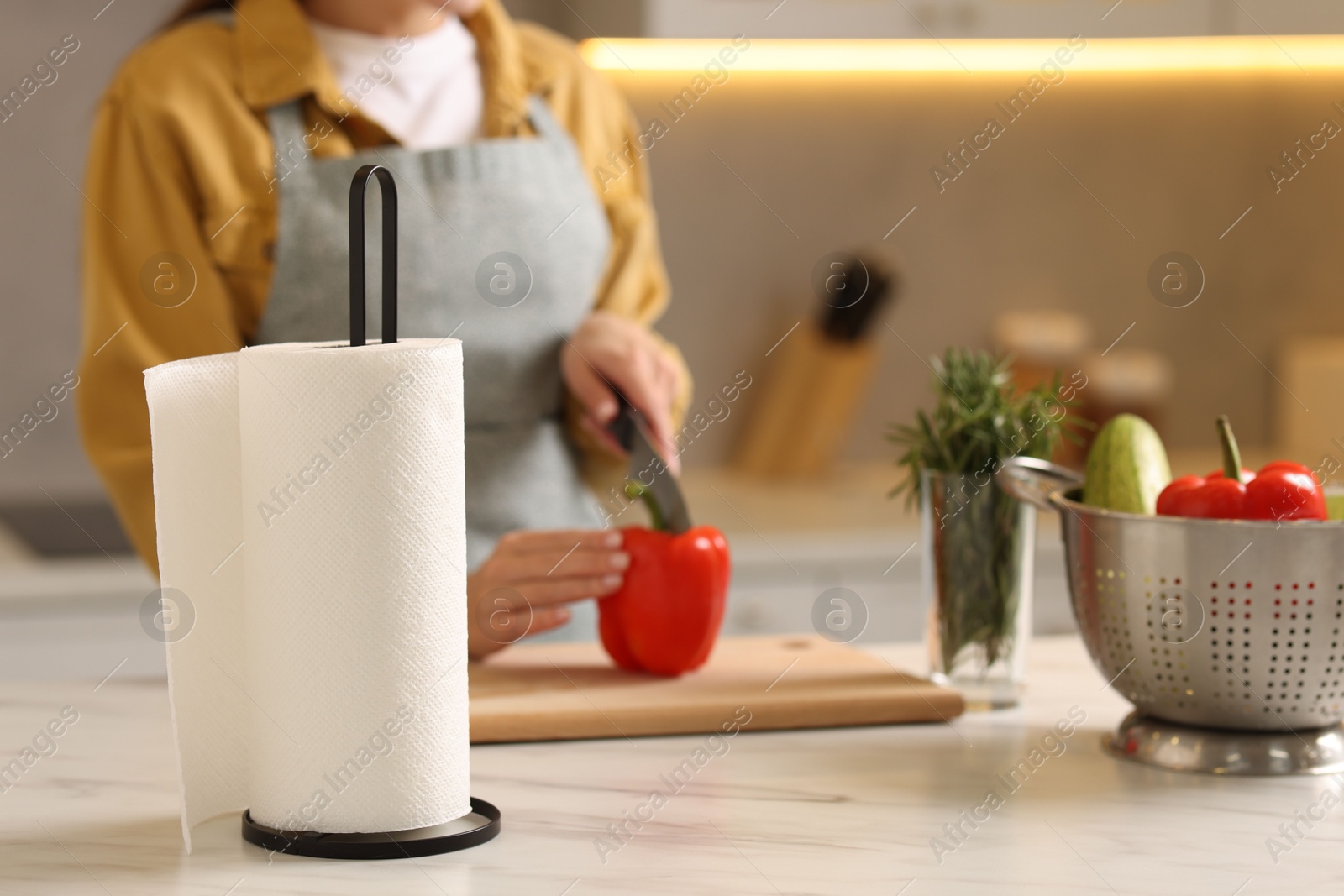 Photo of Woman cutting bell pepper at white marble table in kitchen, focus on roll of paper towels