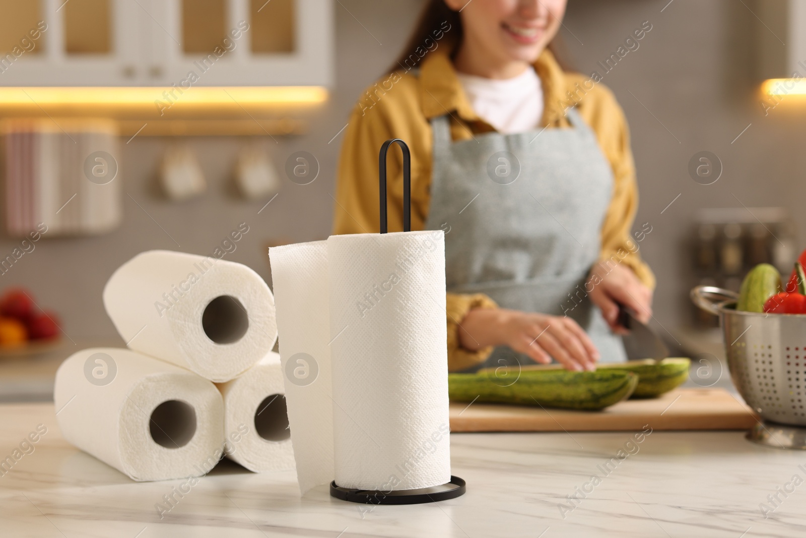 Photo of Woman cutting zucchini at white marble table in kitchen, focus on rolls of paper towels