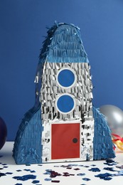 Photo of Bright pinata in shape of spaceship, balloons and confetti on table against blue background