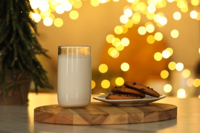 Photo of Glass of milk and tasty chocolate chip cookies on table against blurred Christmas lights. Bokeh effect