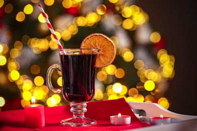 Photo of Aromatic mulled wine in glass cup and burning candles on tray against blurred Christmas lights, bokeh effect