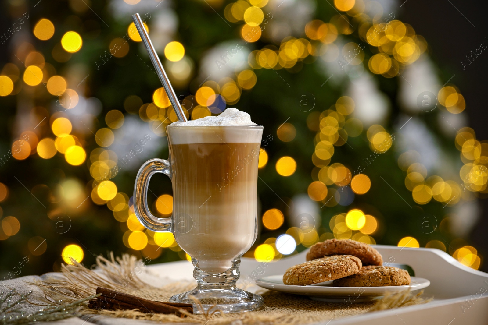 Photo of Aromatic coffee in glass, tasty cookies and cinnamon sticks on tray against blurred Christmas lights, bokeh effect