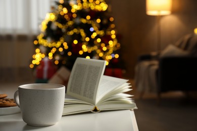 Photo of Cup of hot drink, book and cookies on white table in cozy room decorated for Christmas, closeup. Space for text