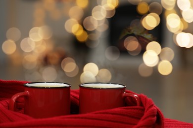 Photo of Tasty cocoa in cups and red scarf against blurred background with lights, closeup. Christmas drink