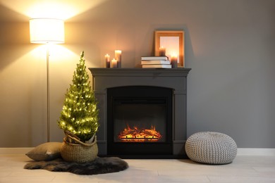 Photo of Small spruce tree in basket decorated with Christmas lights near fireplace indoors