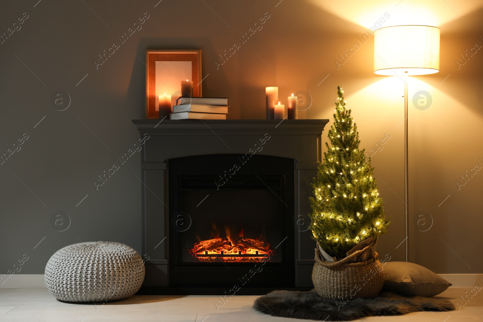 Photo of Small spruce tree in basket decorated with Christmas lights near fireplace indoors