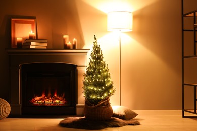 Photo of Small spruce tree in basket decorated with Christmas lights near fireplace indoors