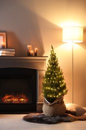 Photo of Small spruce tree in basket decorated with Christmas lights near fireplace indoors