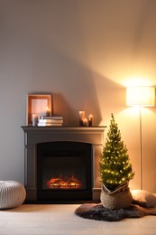 Photo of Small spruce tree in basket decorated with Christmas lights near fireplace indoors