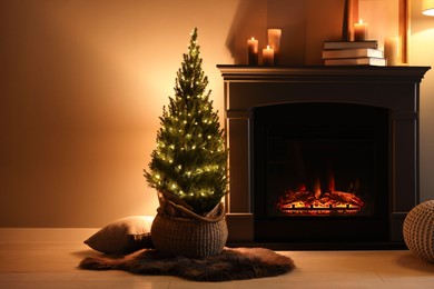 Photo of Small spruce tree in basket decorated with Christmas lights near fireplace indoors