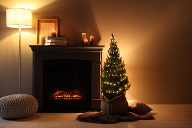 Photo of Small spruce tree in basket decorated with Christmas lights near fireplace indoors