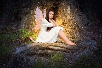 Image of Charming fairy sitting on tree root. Mesmerizing girl with ethereal wings in forest