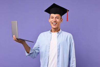 Happy student with laptop after graduation on violet background