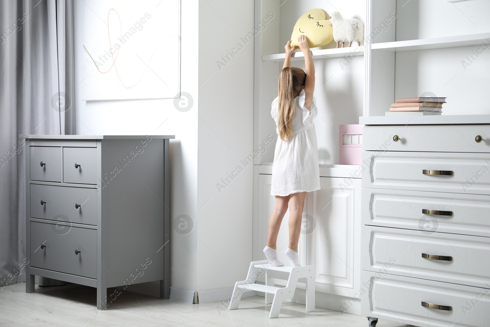 Photo of Little girl standing on step stool and reaching for toys on shelf indoors, back view