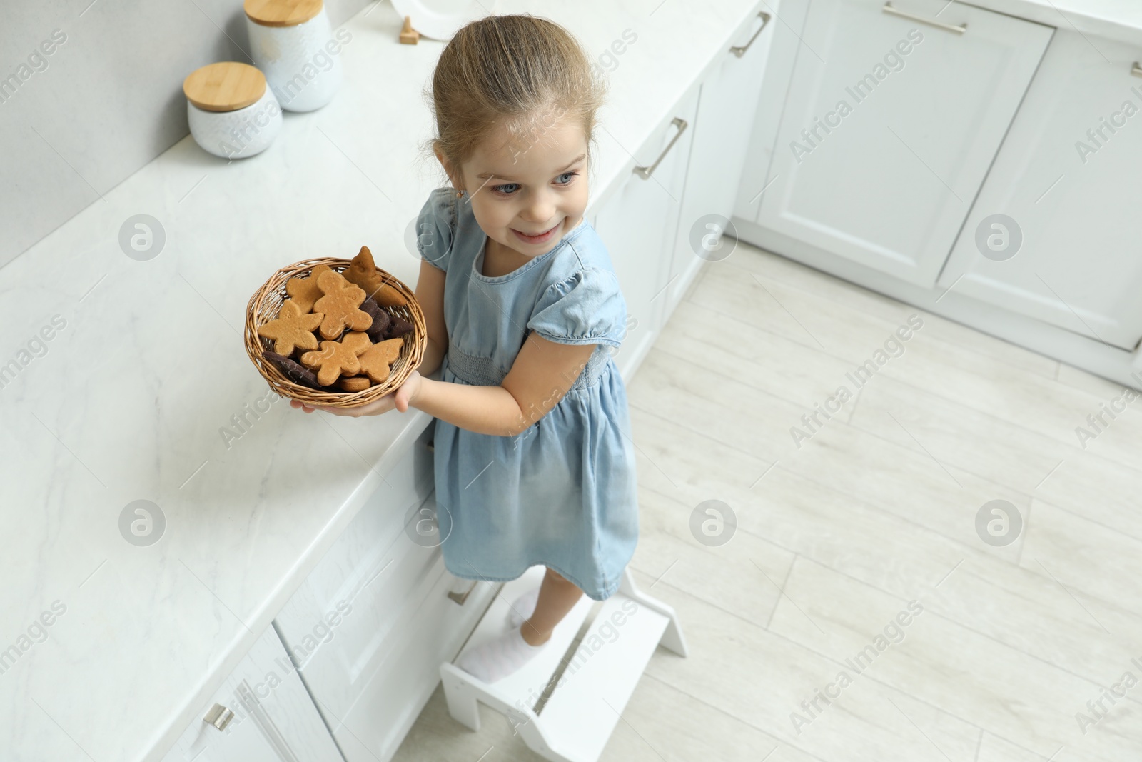 Photo of Little girl standing on step stool with bowl of cookies in kitchen, above view. Space for text