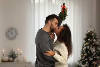 Happy couple kissing under mistletoe bunch in room decorated for Christmas