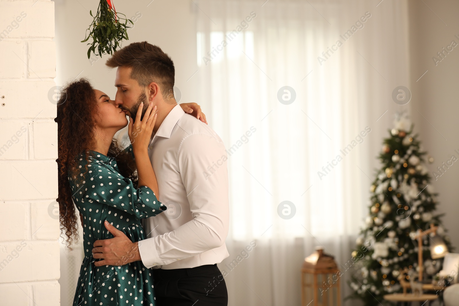 Photo of Happy couple kissing under mistletoe bunch in room decorated for Christmas