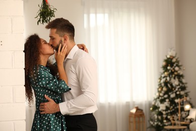 Photo of Happy couple kissing under mistletoe bunch in room decorated for Christmas