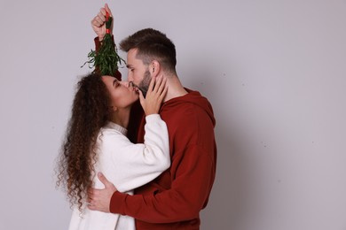 Photo of Happy couple kissing under mistletoe bunch on light grey background. Space for text