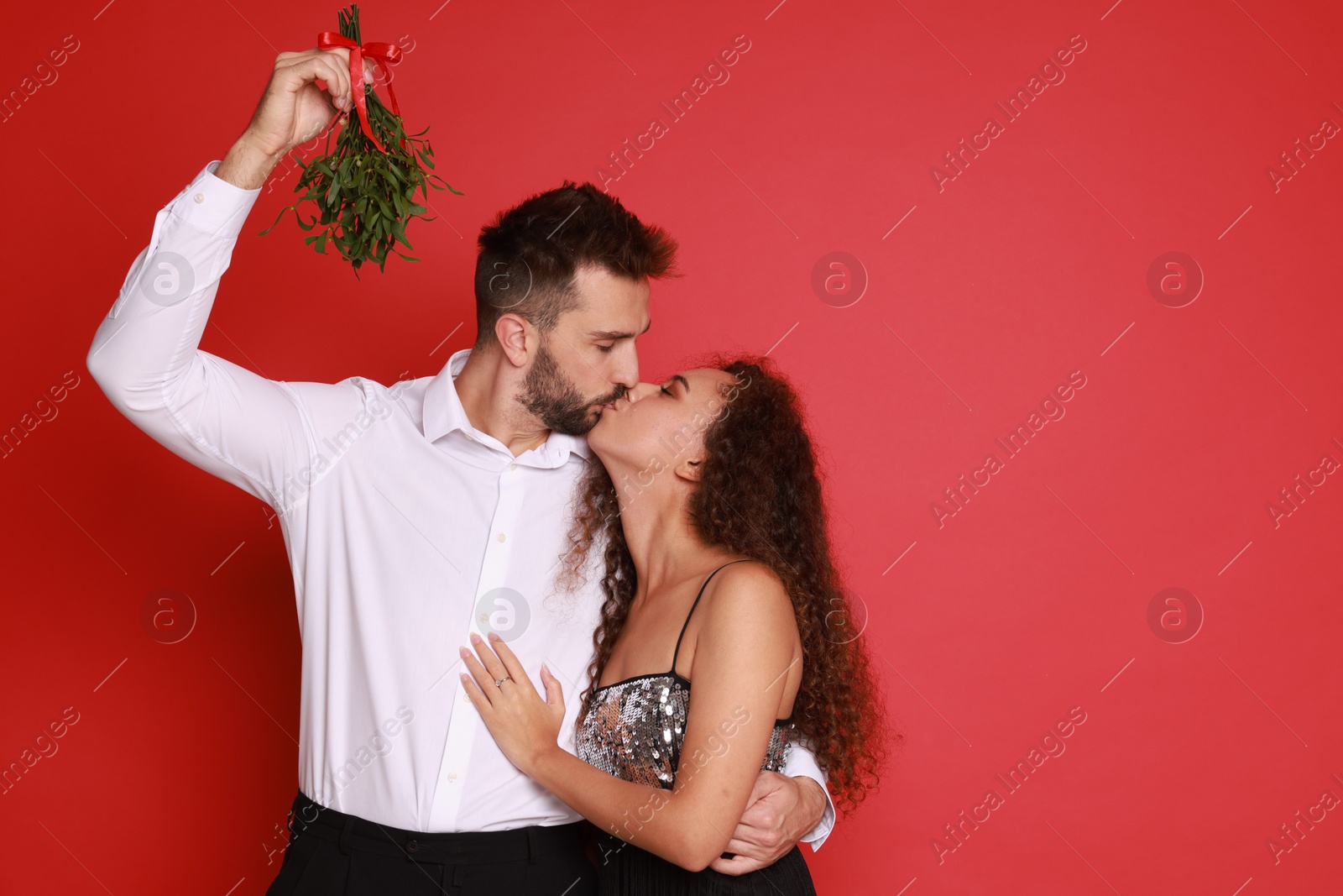 Photo of Happy couple kissing under mistletoe bunch on red background. Space for text