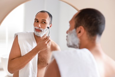 Photo of Handsome man shaving with razor near mirror in bathroom