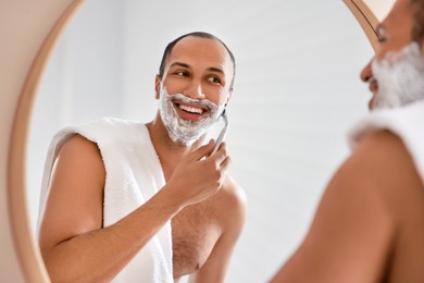 Photo of Smiling man shaving with razor near mirror in bathroom