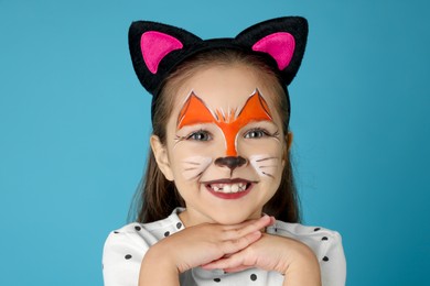 Photo of Cute girl with painted face and ears as cat on light blue background