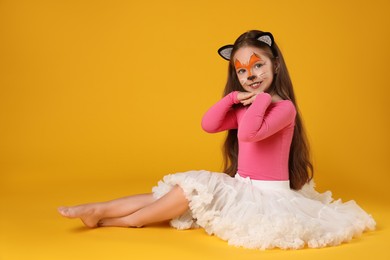 Photo of Cute girl with painted face and ears as cat on orange background, space for text