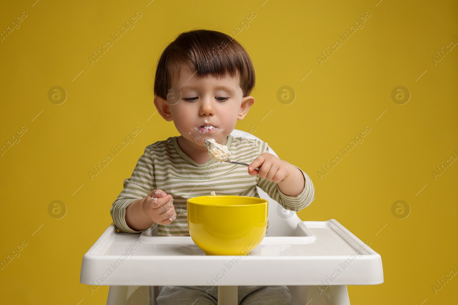 Photo of Cute little kid eating healthy baby food from bowl in high chair on yellow background