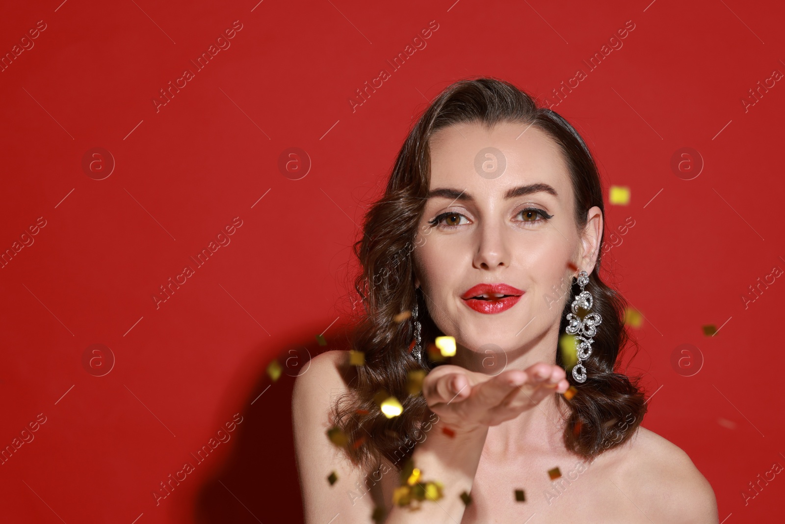 Photo of Woman with perfect makeup blowing confetti on red background, space for text. Christmas celebration