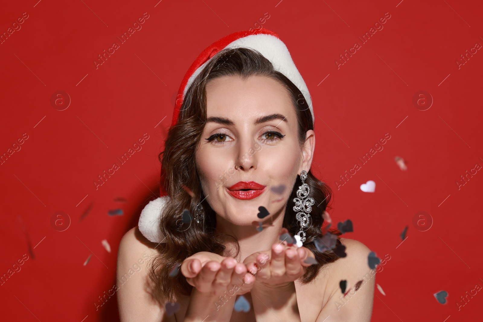 Photo of Woman with perfect makeup in Santa hat blowing confetti on red background. Christmas celebration