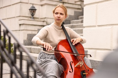 Beautiful young woman playing cello on stairs outdoors. Classic musical instrument