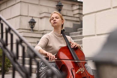 Photo of Beautiful young woman playing cello on stairs outdoors. Classic musical instrument